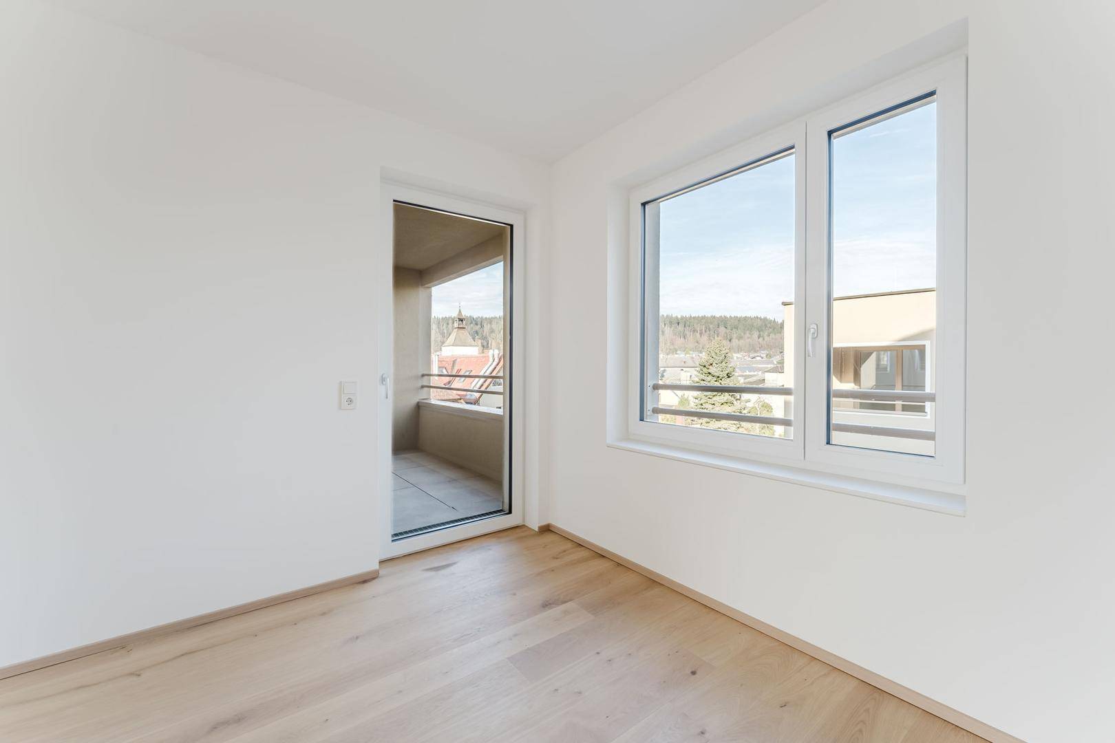 Ruhiges Zimmer mit Holzboden, Blick ins Grüne und Zugang zu einem überdachten Balkon.