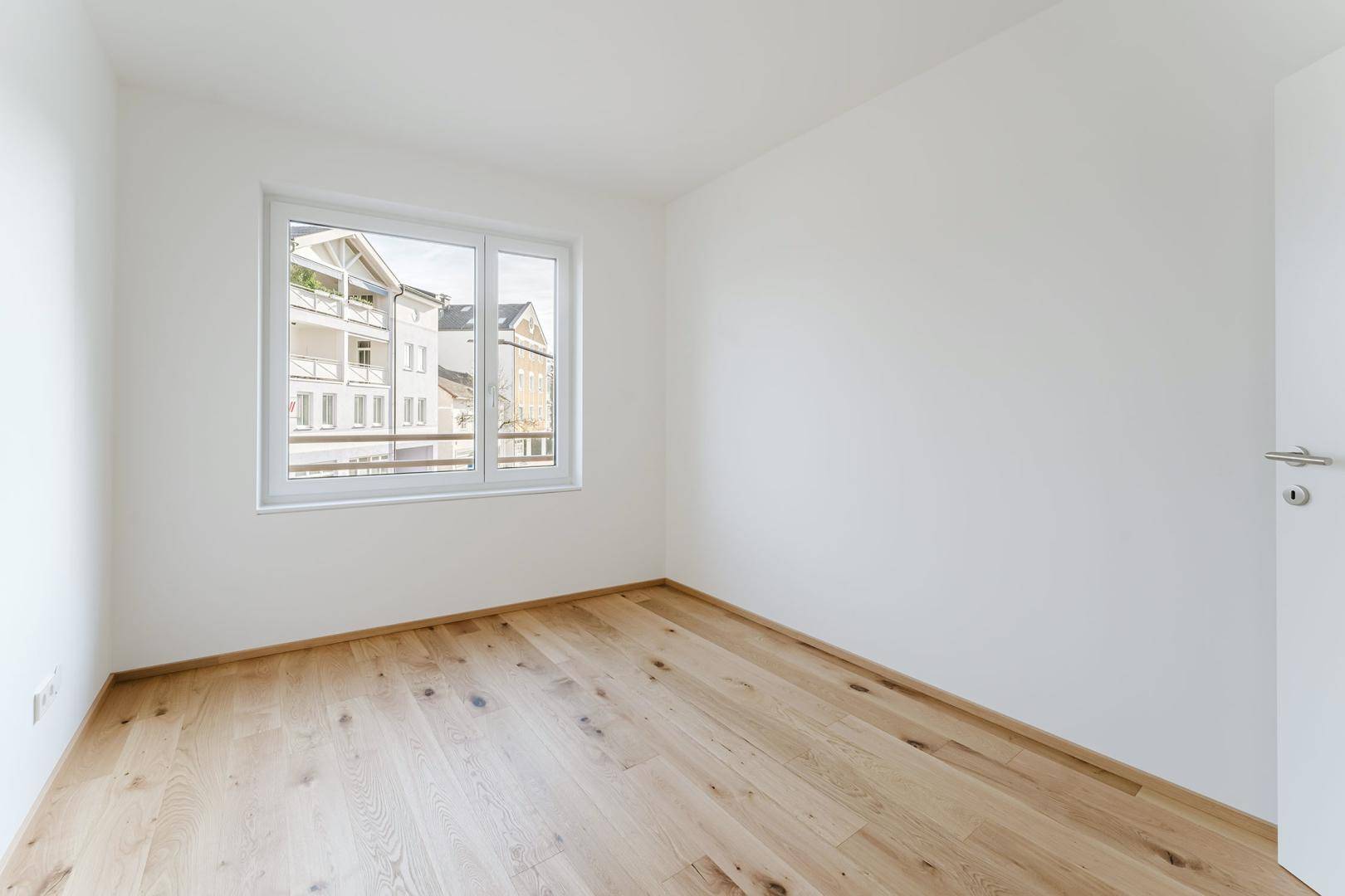 Helles, leeres Schlafzimmer mit Holzboden und Blick auf die Nachbargebäude.