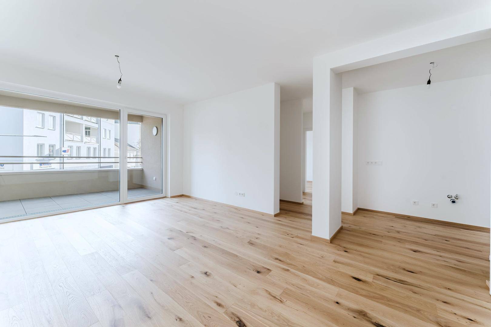 Helles, leeres Wohnzimmer mit Holzboden und großem Schiebefenster zum Balkon.
