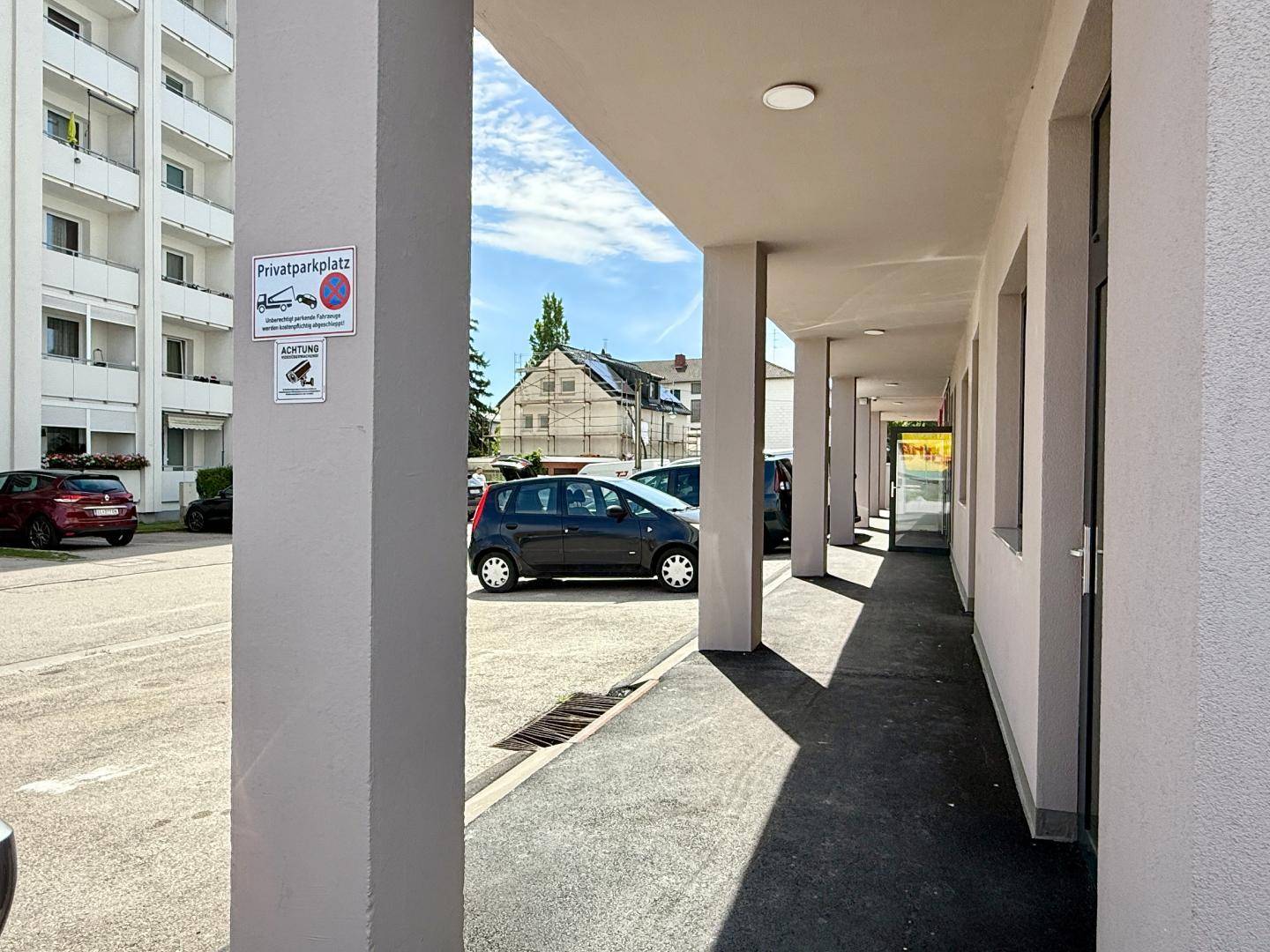 Außenansicht eines Gebäudes mit überdachtem Gehweg und einem angrenzenden Privatparkplatz.