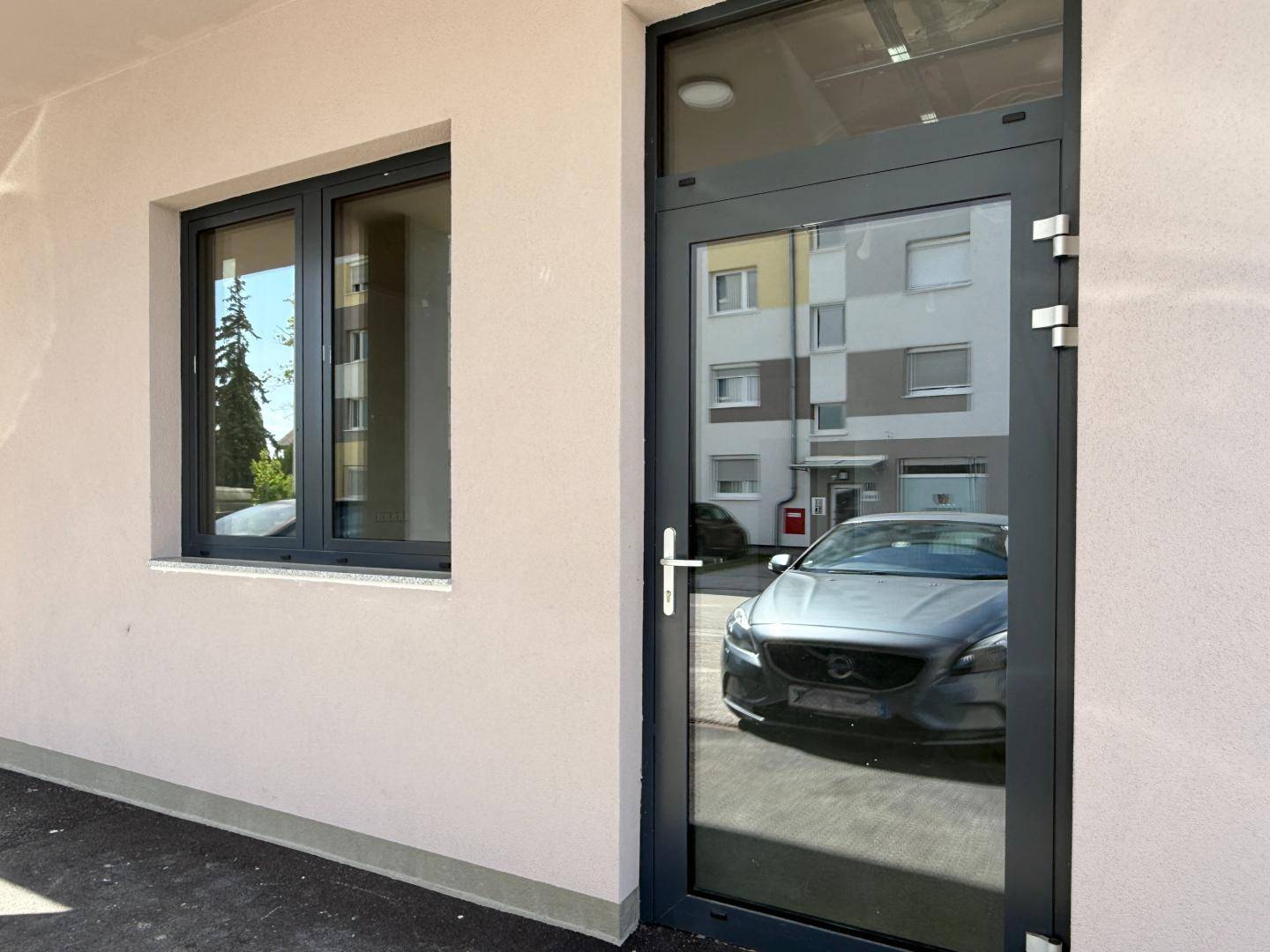 Eingangsbereich einer Gewerbefläche mit großer Glastür und Fenster, reflektiert parkende Autos.