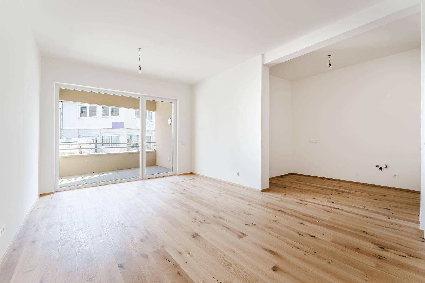 Leerer, heller Wohnbereich mit Holzboden, weißen Wänden und großem Balkonzugang.