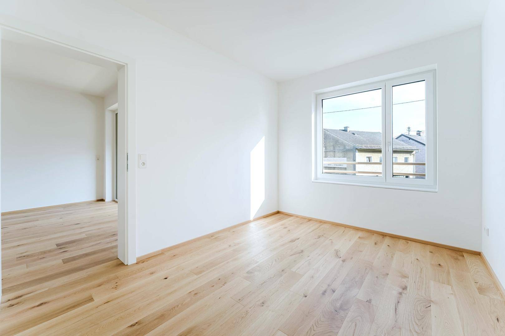 Leeres Schlafzimmer mit hellem Holzfußboden und Fenster, das einen Blick nach draußen bietet.