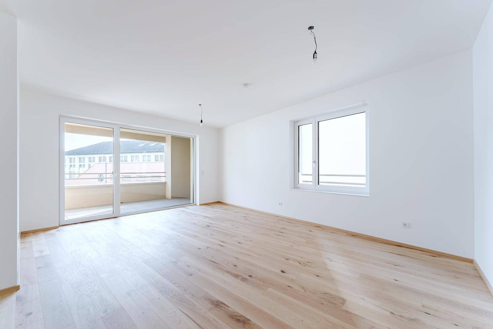 Geräumiges, leeres Zimmer mit hellem Holzfußboden und großem Schiebetürzugang zum Balkon.