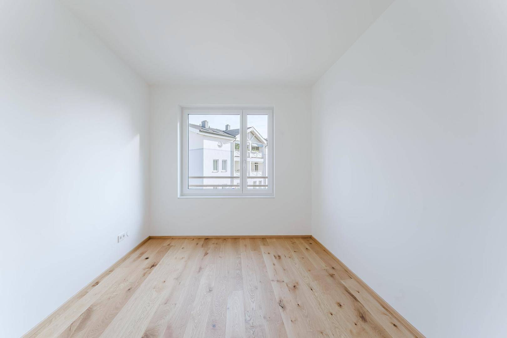 Leeres Zimmer mit hellem Holzboden und einem Fenster, das viel Tageslicht hereinlässt.
