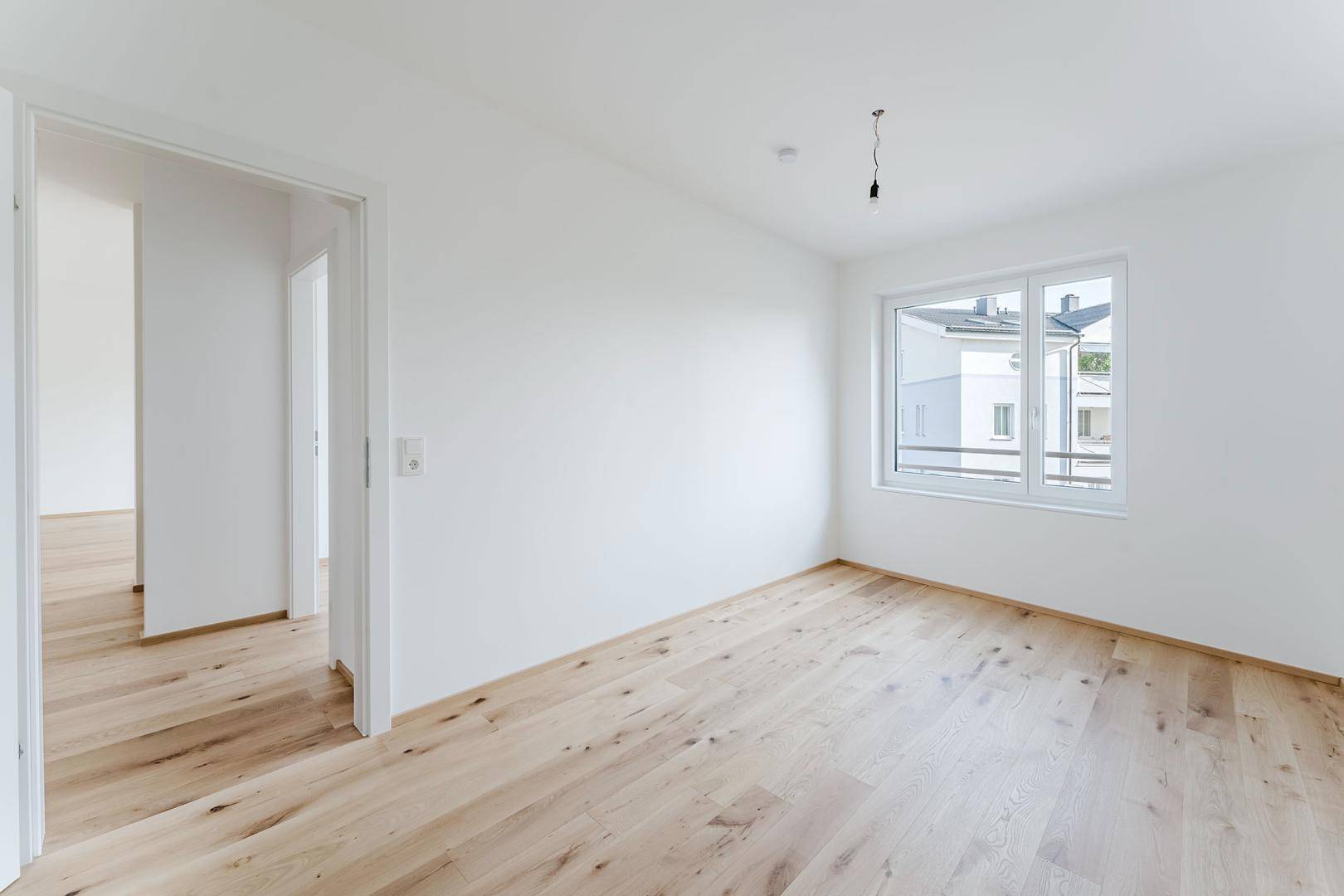 Helles, leeres Zimmer mit Holzboden und einem Fenster, das den Blick auf die Umgebung freigibt.