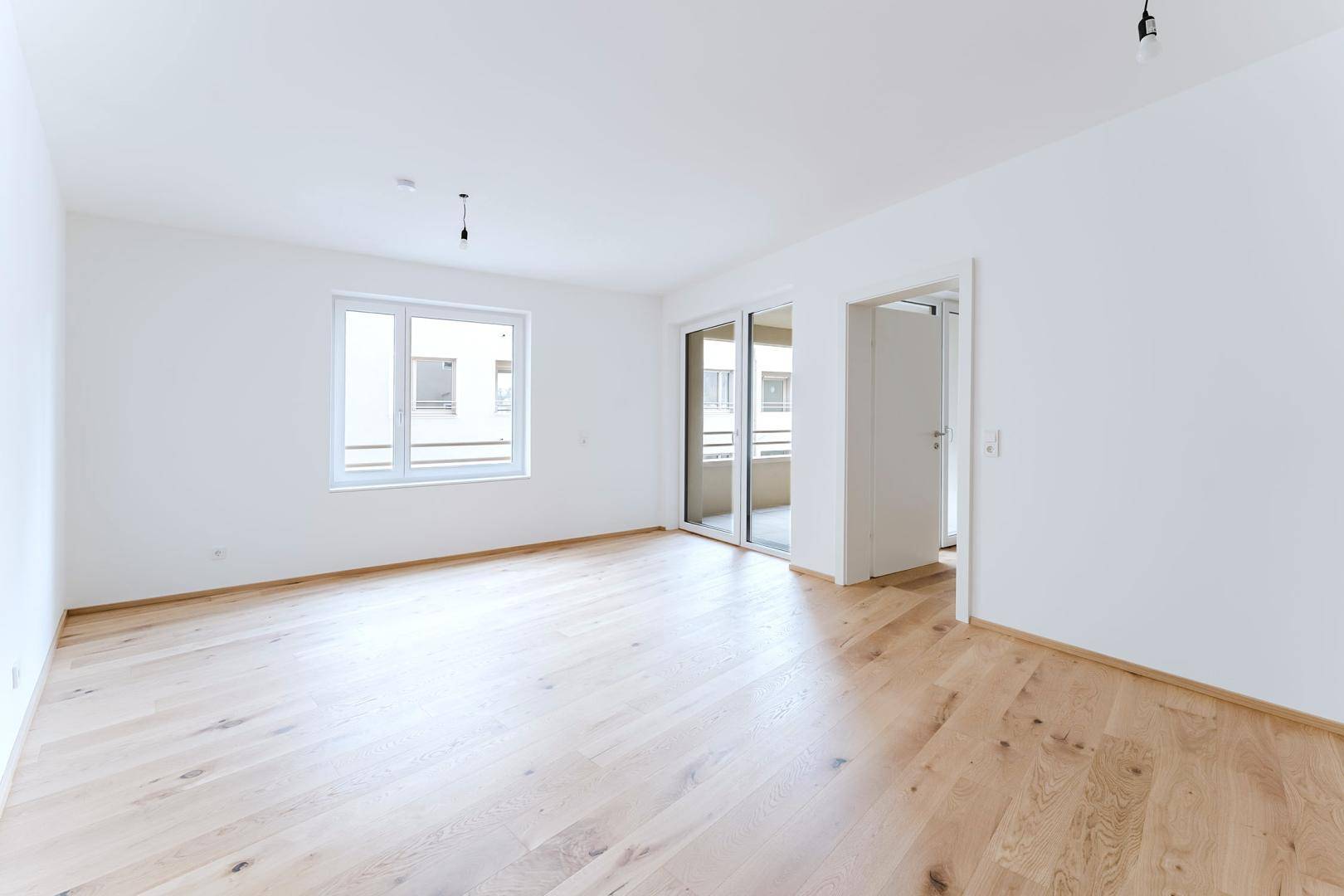 Heller, leerer Wohnraum mit großen Fenstern, Holzboden und Zugang zu einem Außenbereich.