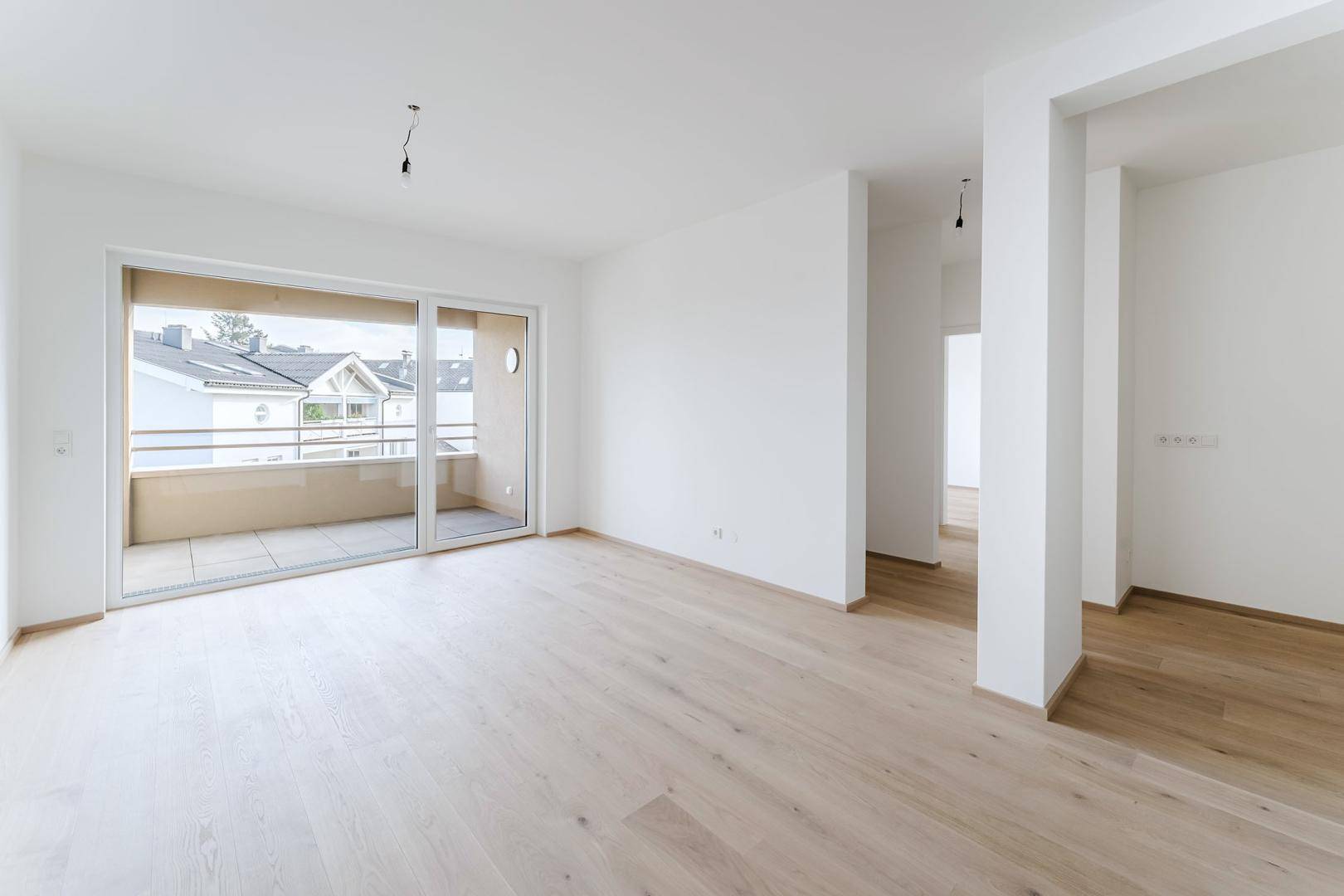 Heller, leerer Wohnraum mit Holzboden und großem Schiebefenster zum Balkon.