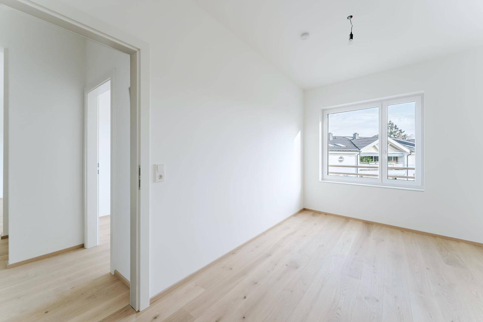 Leeres Zimmer mit hellem Holzboden und Fenster, das viel Tageslicht hereinlässt.