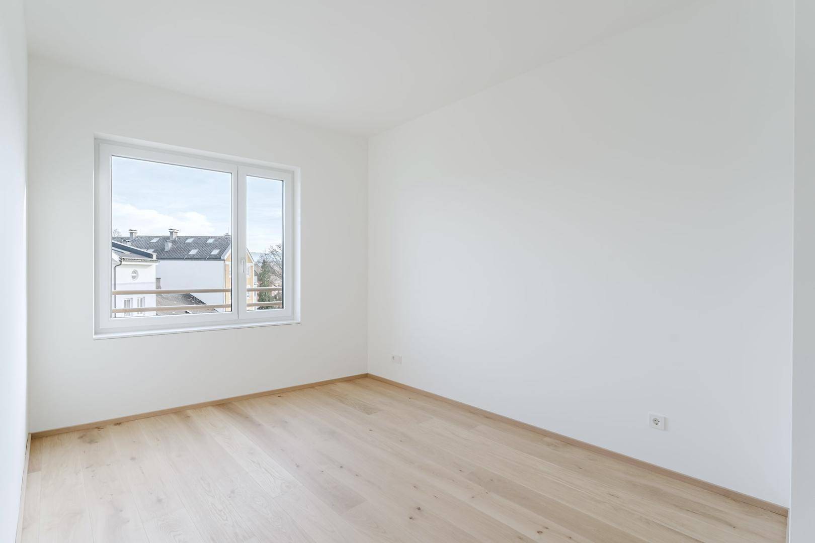Helles, unmöbliertes Zimmer mit Holzboden und großem Fenster mit Blick nach draußen.