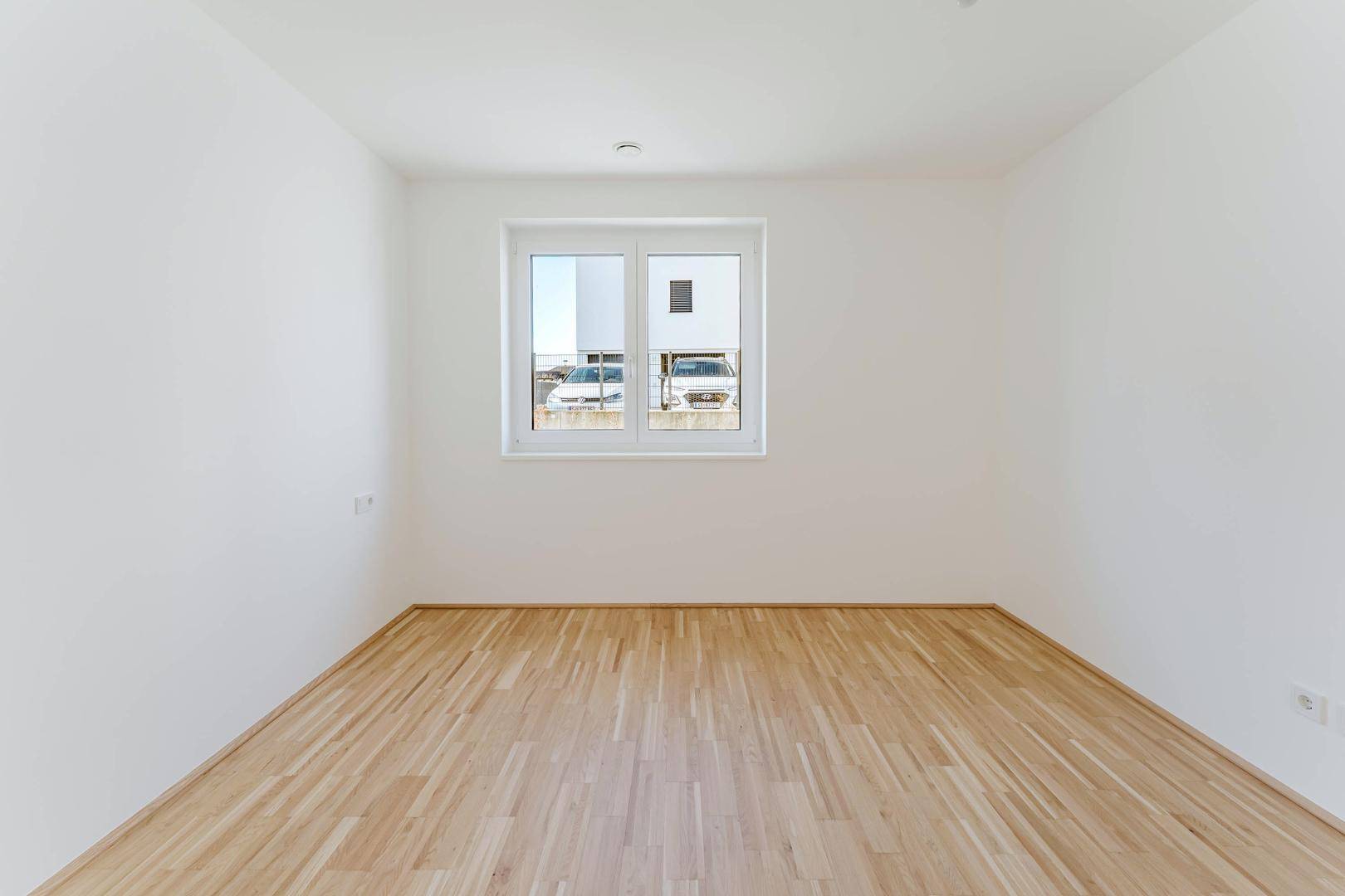 Leeres Zimmer mit Holzboden und großem Fenster, das viel Tageslicht in den Raum bringt.