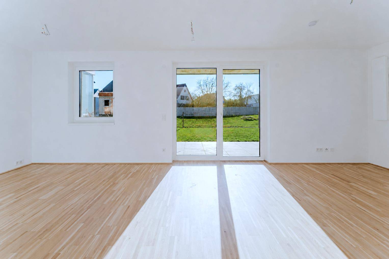 Heller, leerer Raum mit Holzboden und großen Fenstern, die viel Tageslicht hereinlassen.