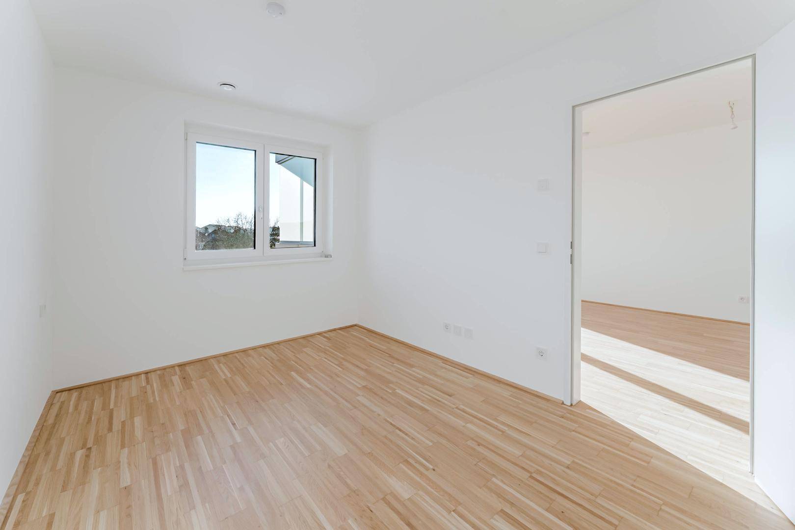 Helles Zimmer mit Parkettboden und großem Fenster, ideal als Schlaf- oder Arbeitszimmer.