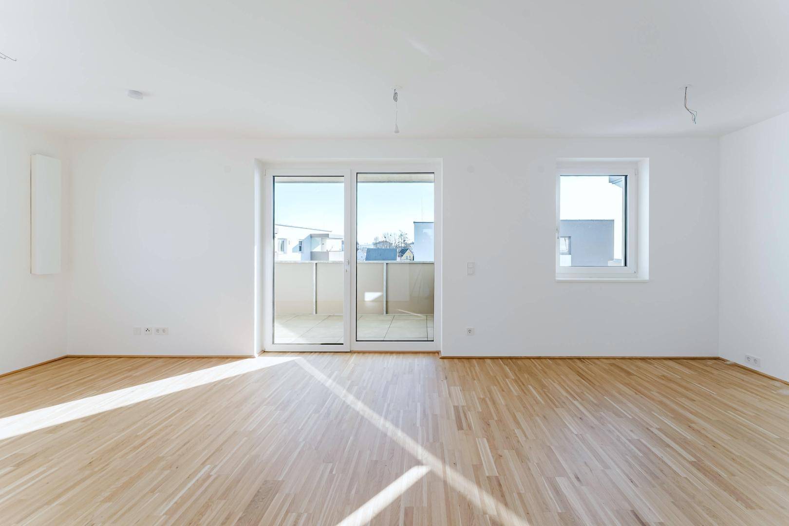 Heller, leerer Wohnraum mit hellem Parkettboden und Zugang zu einem sonnigen Balkon.