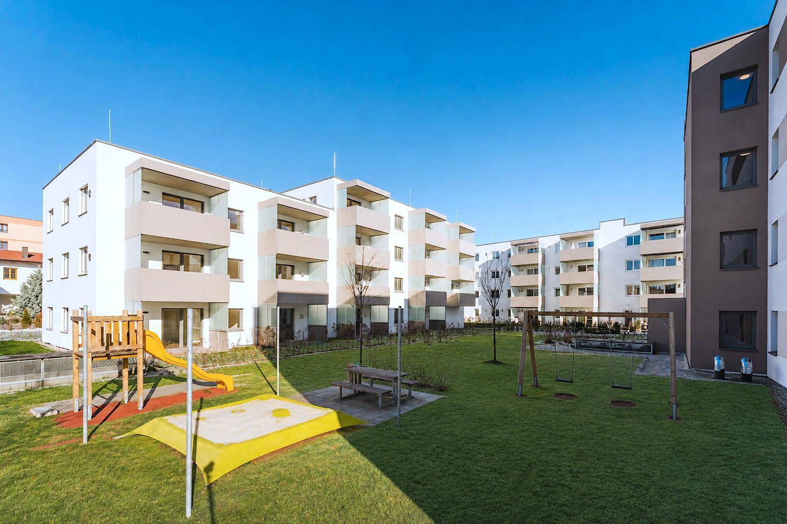 Weitläufige Außenanlage mit mehreren modernen Wohngebäuden, Rasenflächen und Spielplatz.