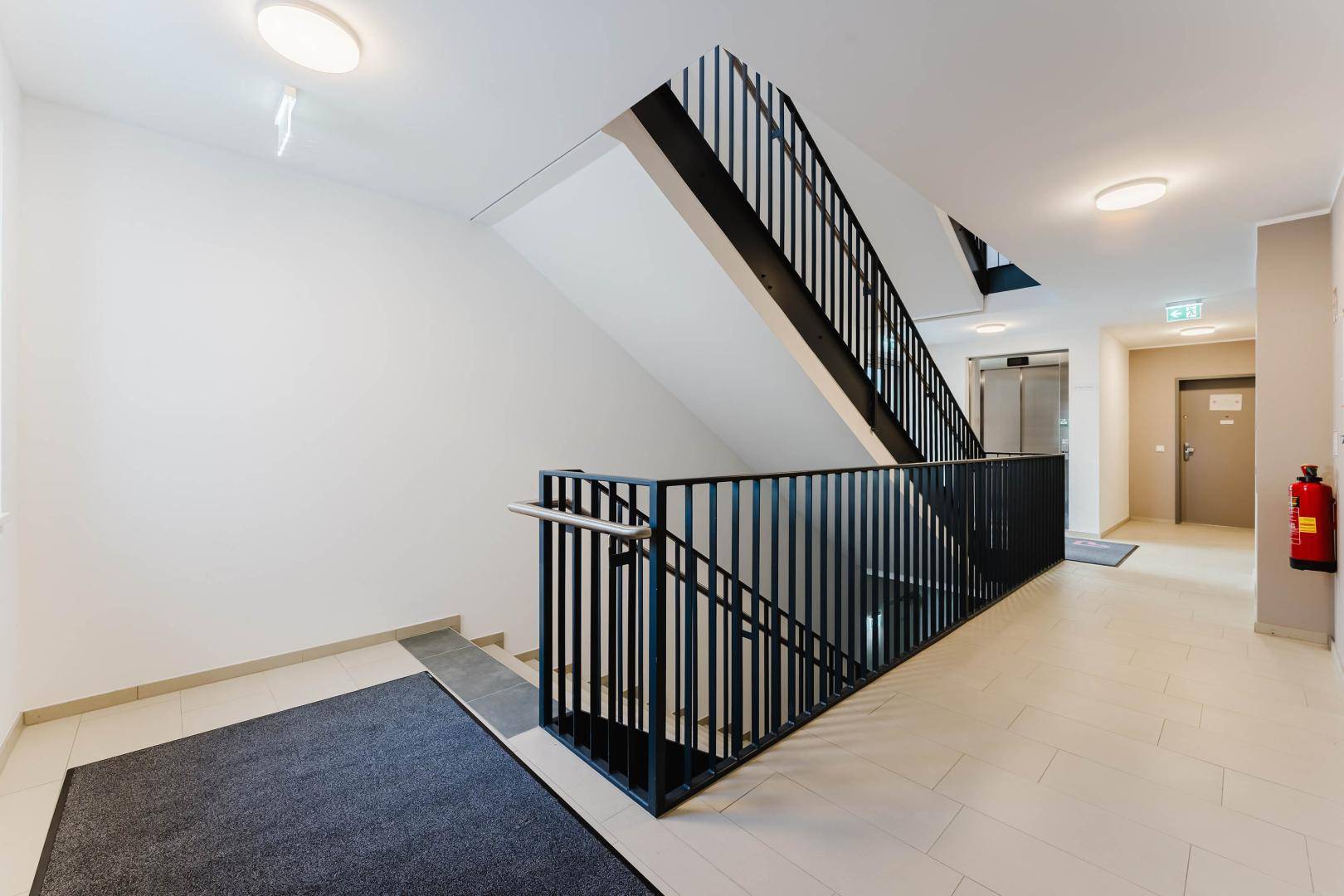 Modernes Treppenhaus mit schwarzem Geländer und einem Aufzug im Hintergrund.