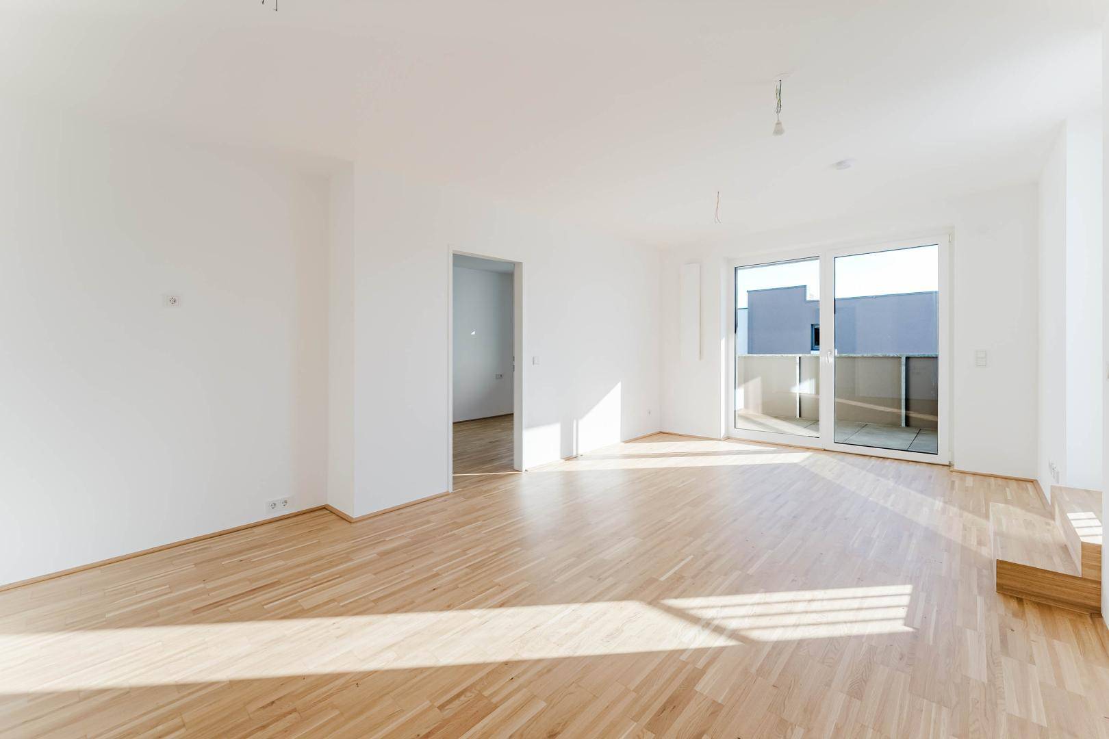 Lichtdurchfluteter Raum mit Parkettboden und Zugang zu einem Balkon oder einer Terrasse.