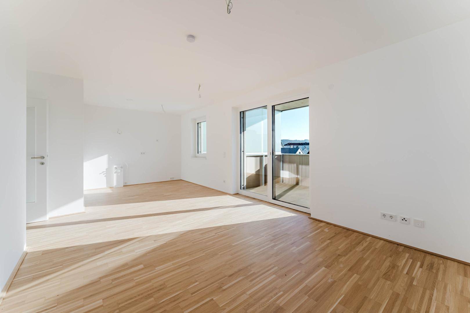 Heller, leerer Wohnbereich mit Parkettboden und Zugang zu einem Balkon in einem Neubau.