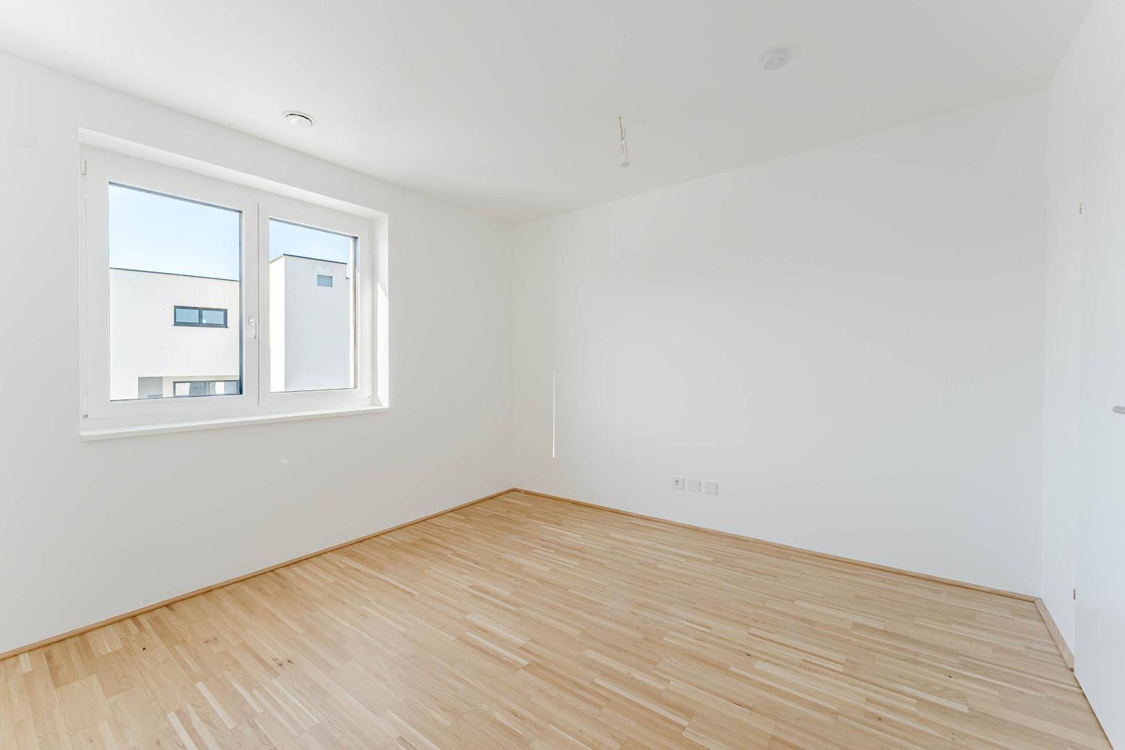 Helles Schlafzimmer mit Parkettboden und großem Fenster in einem modernen Neubau.