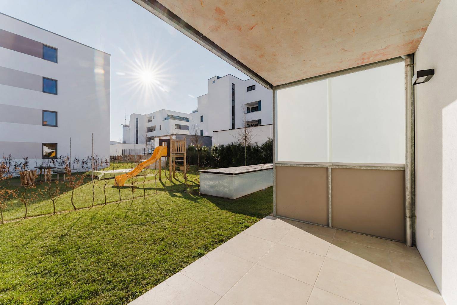 Sonniger Terrassenbereich mit Fliesenboden und Blick auf den Gemeinschaftsgarten mit Spielplatz.