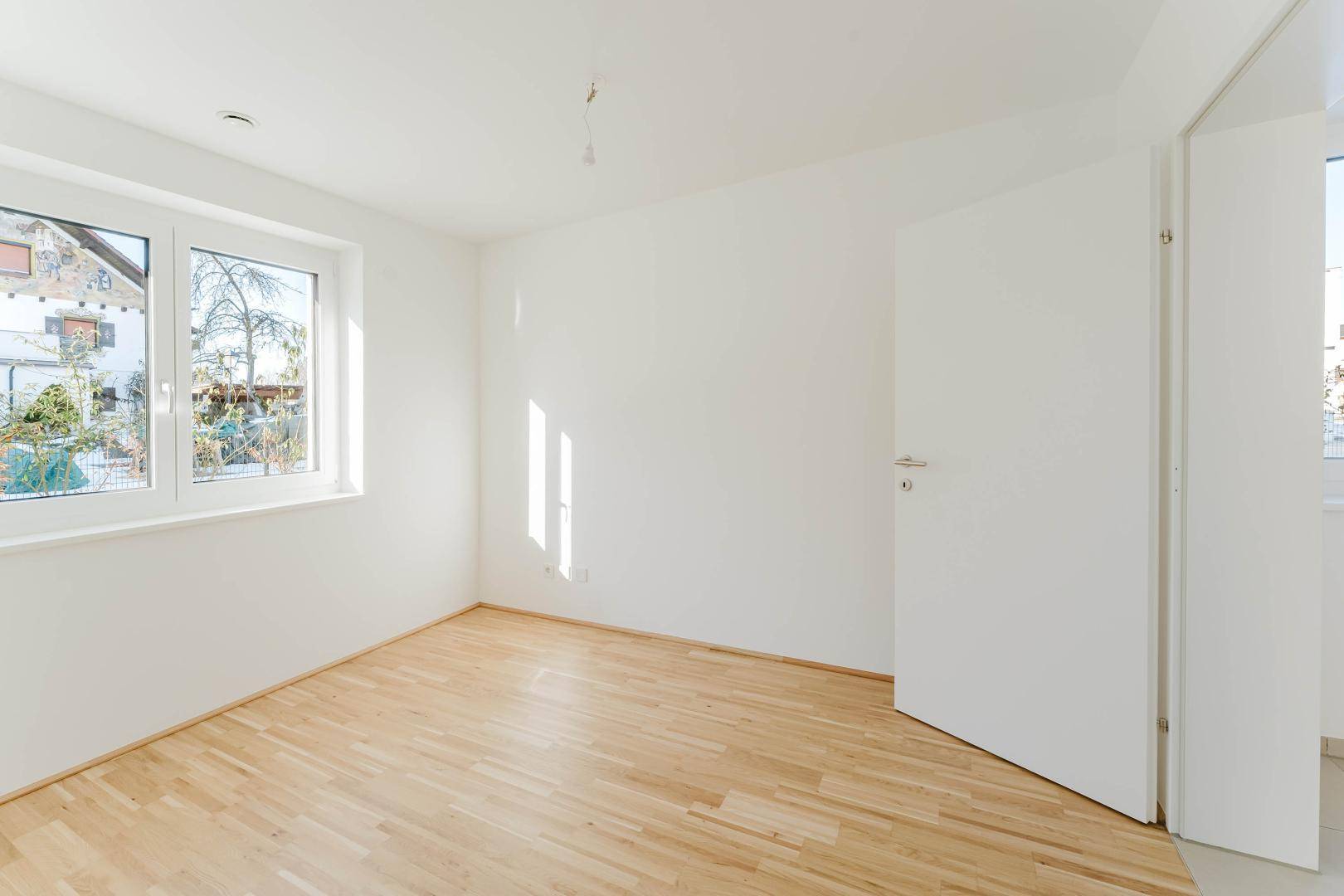 Helles Schlafzimmer mit Parkettboden und großem Fenster, das viel Tageslicht hereinlässt.