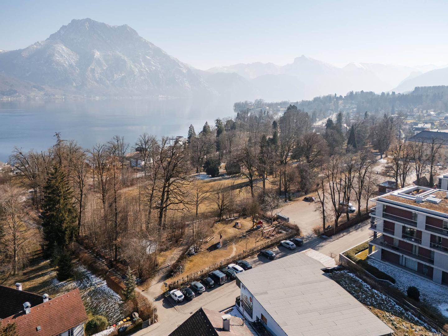 Landschaftsbild mit See, majestätischen Bergen und einem Spielplatz, umgeben von kahlen Bäumen.