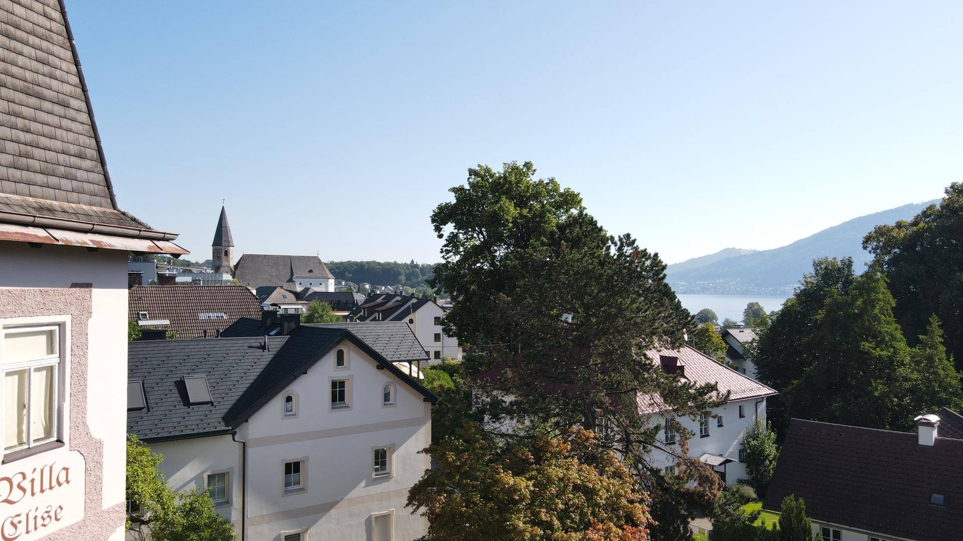 Ausblick über Dächer, einen Kirchturm und Bäume mit einem See in der Ferne unter blauem Himmel.