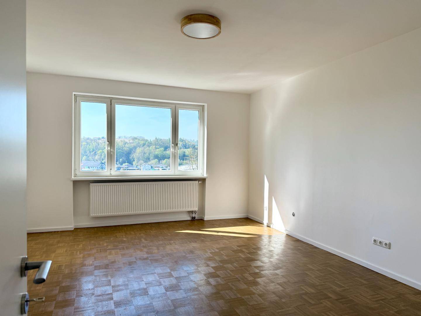 Leeres, helles Wohnzimmer mit Parkettboden und großem Fenster mit Weitblick.