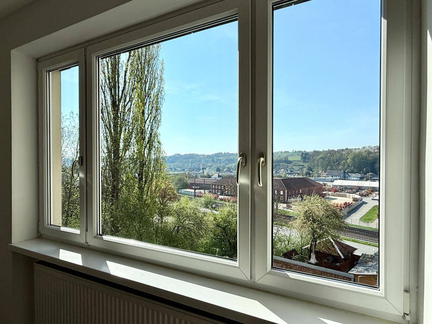 Fensterblick aus dem Wohnzimmer auf eine grüne Landschaft mit Gebäuden und Bäumen.