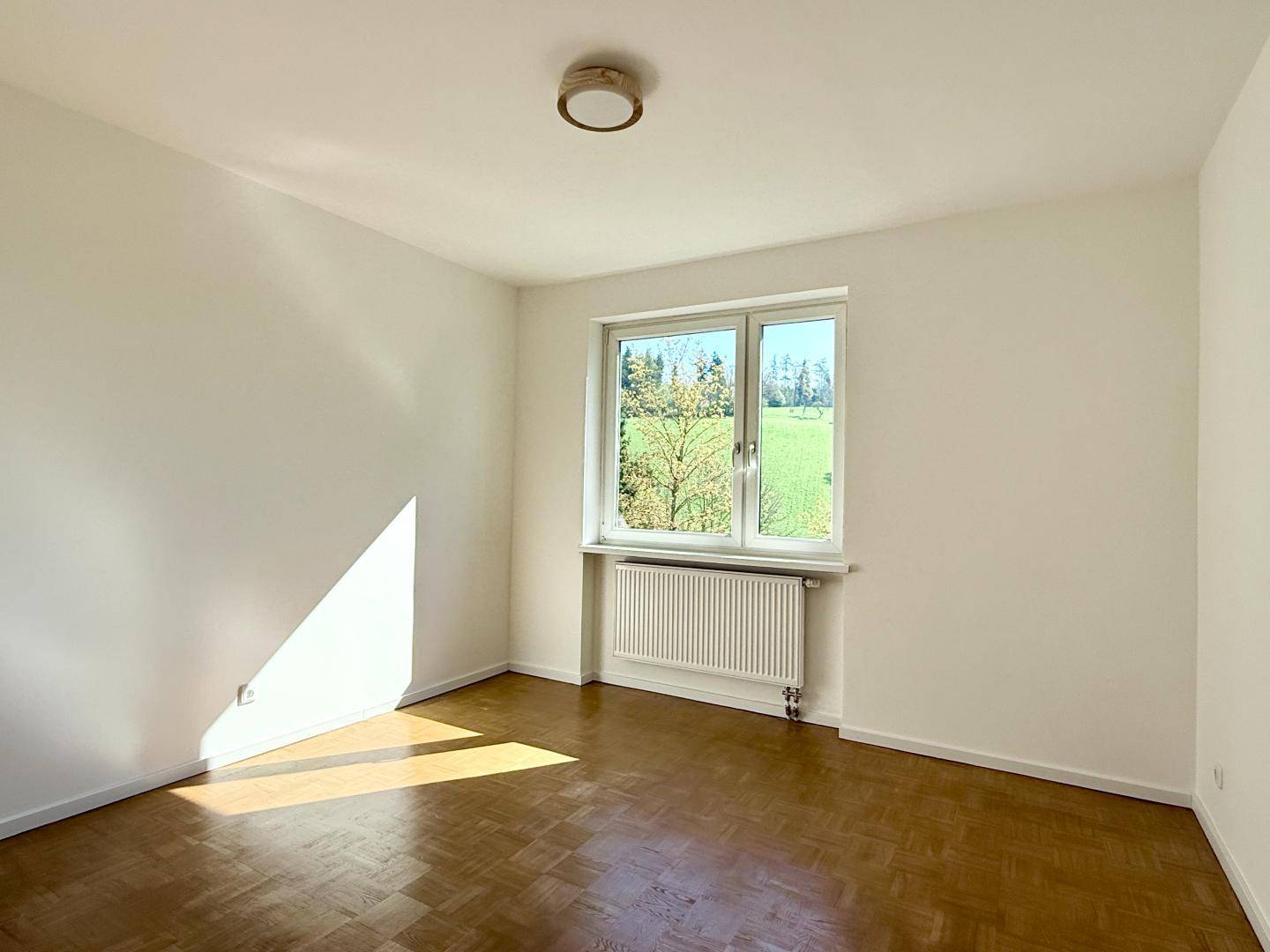 Helles Schlafzimmer mit Parkettboden und Fenster mit Blick auf grüne Felder.