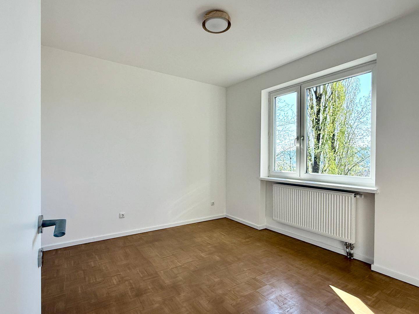 Leeres Schlafzimmer mit Parkettboden und Fenster, das viel Tageslicht hereinlässt.