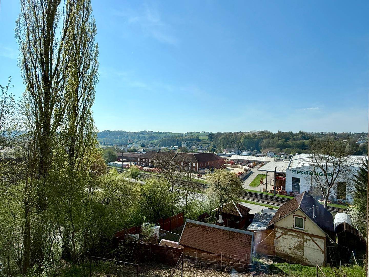 Weitblick über eine grüne Landschaft mit Bäumen, Gebäuden und einem klaren blauen Himmel.
