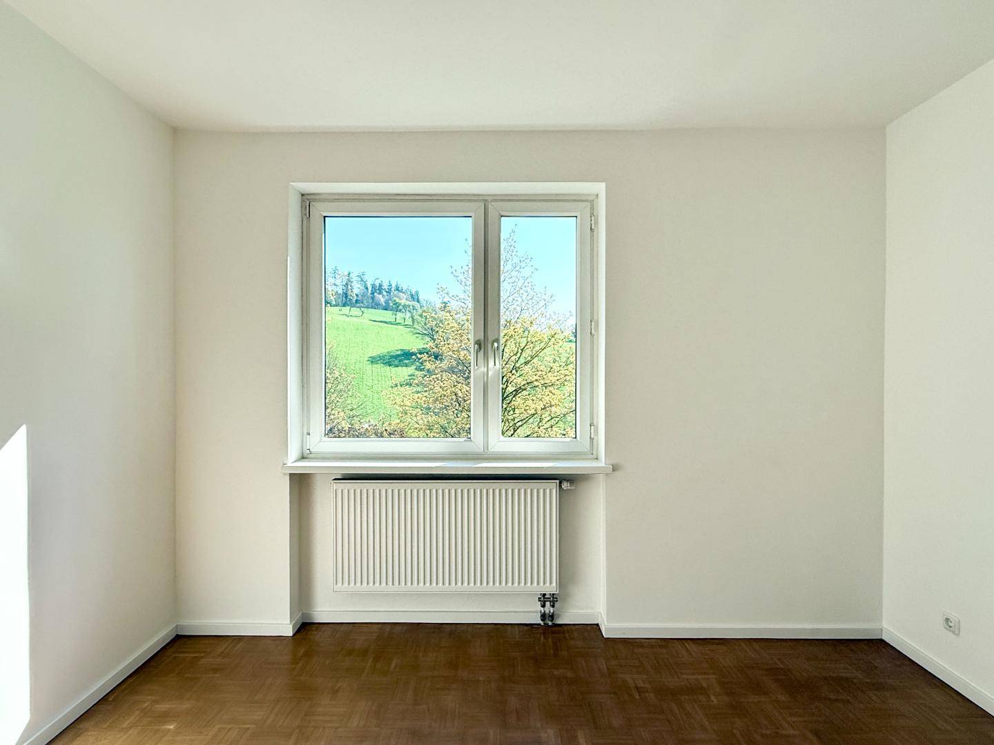 Unmöbliertes Schlafzimmer mit Parkettboden und großem Fenster mit Blick ins Grüne.