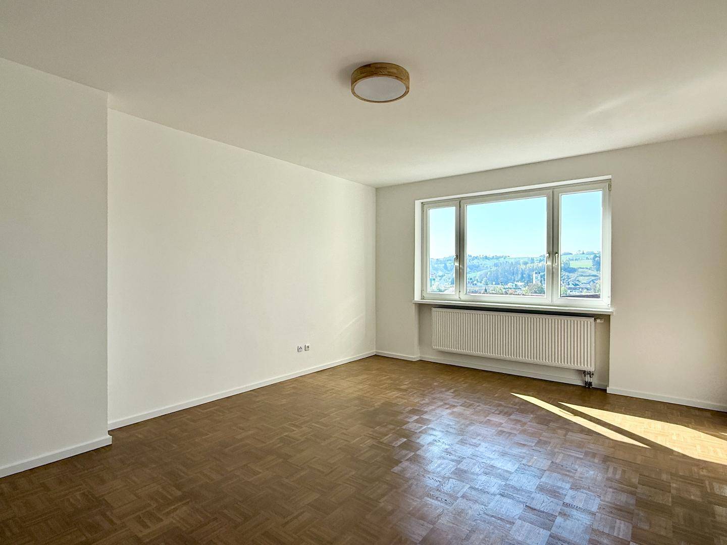Geräumiges, leeres Wohnzimmer mit Parkettboden und Fenster, das viel Tageslicht hereinlässt.
