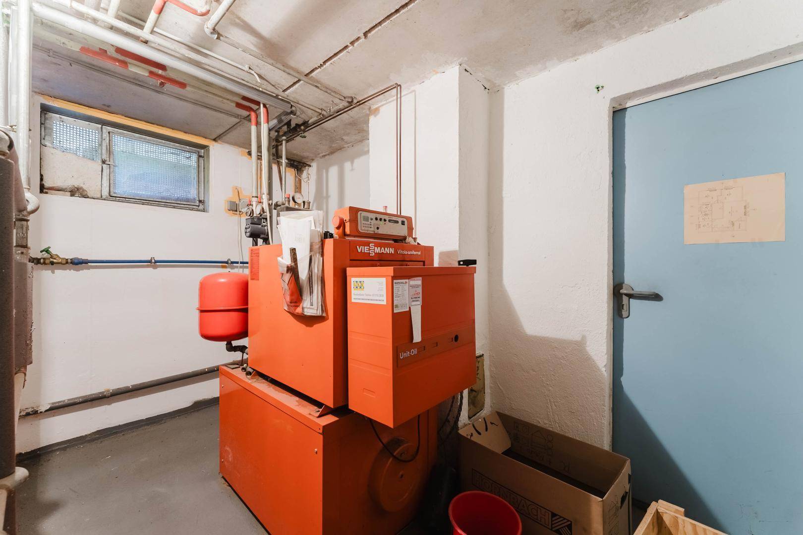 Heizungsraum im Keller mit orangefarbener Heizungsanlage und sichtbaren Rohren.