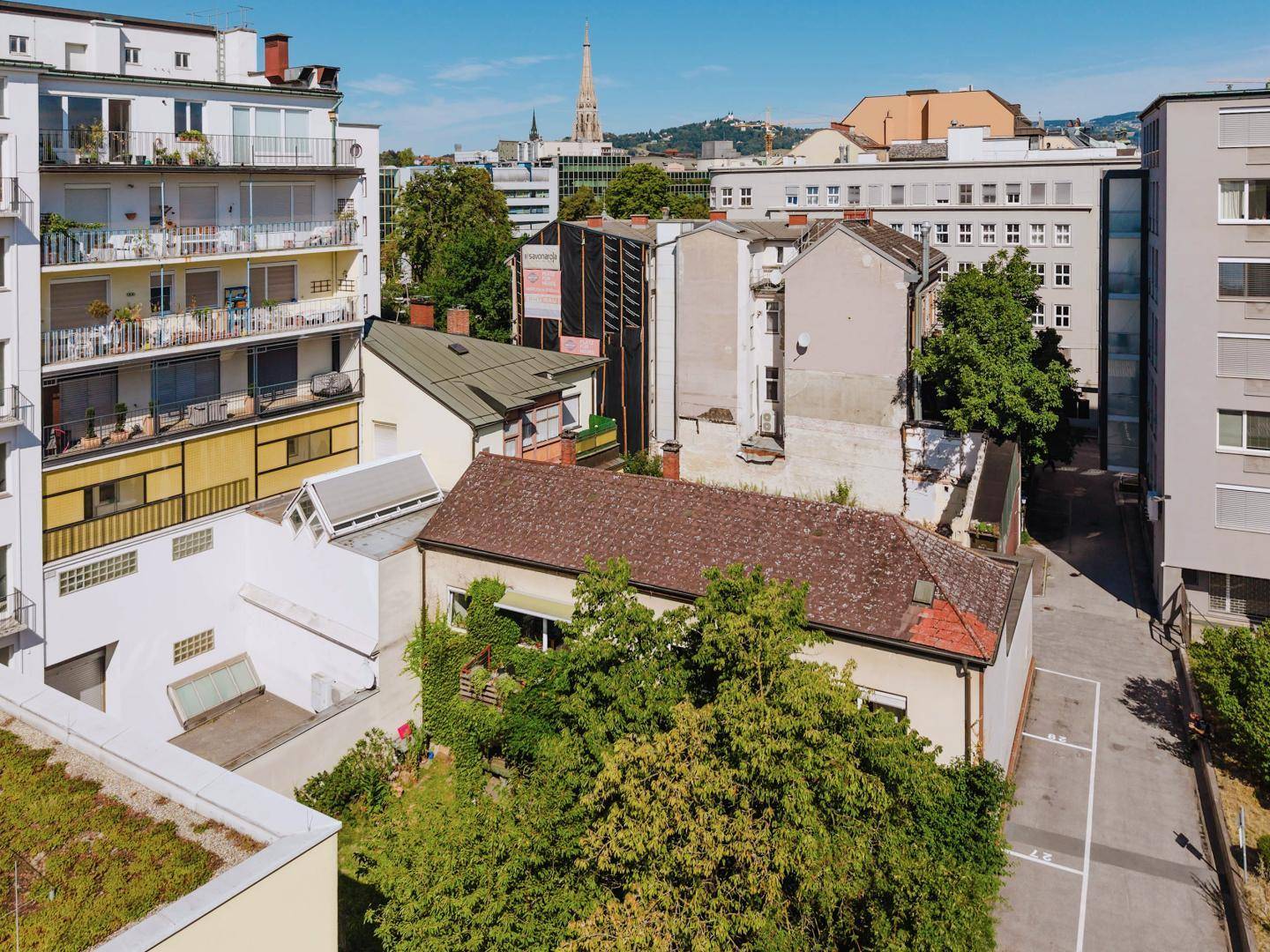 Luftaufnahme von Gebäuden in einer Stadt, darunter ein älteres Haus mit rotem Ziegeldach und Kirchturm in der Ferne.