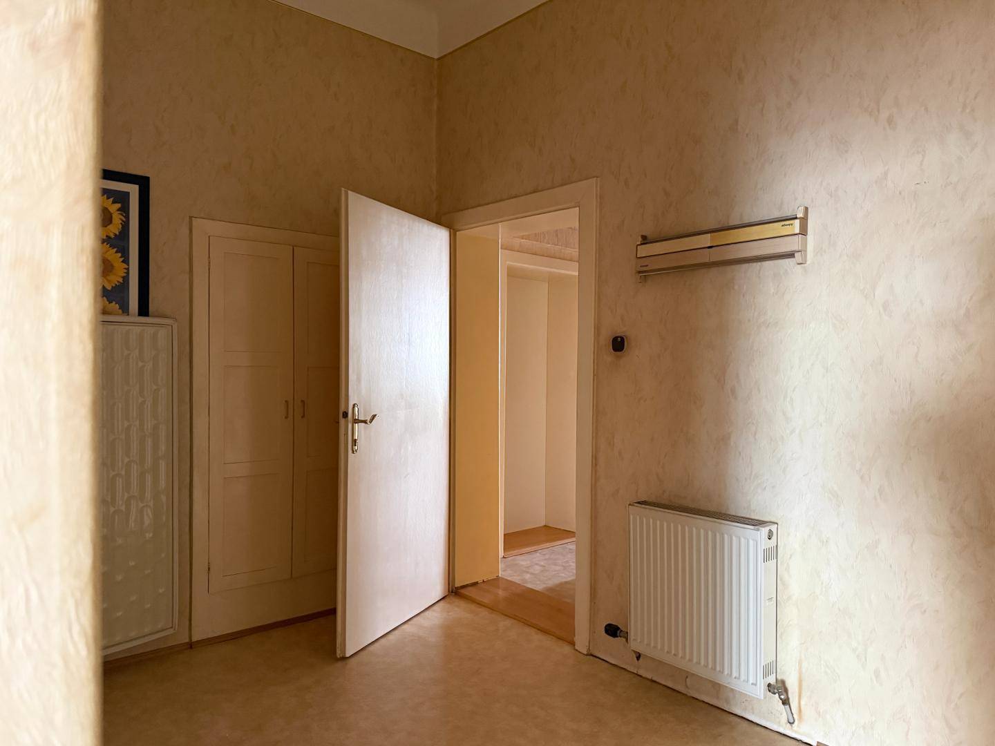 Raum mit heller Tapete, einer offenen Tür, die in einen weiteren Raum führt, und einem Heizkörper.