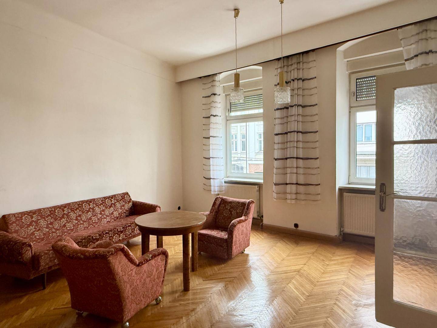 Geräumiges Wohnzimmer mit Fischgrätparkett, Vintage-Möbeln und großen Fenstern, die viel Licht hereinlassen.