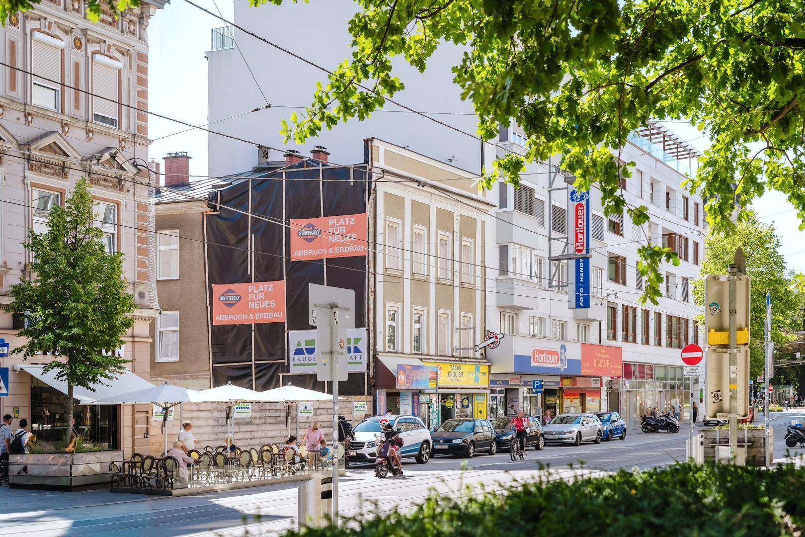 Eine sonnige Stadtstraße mit Gebäuden, einem Außenbereich mit Sitzgelegenheiten und grünen Bäumen.