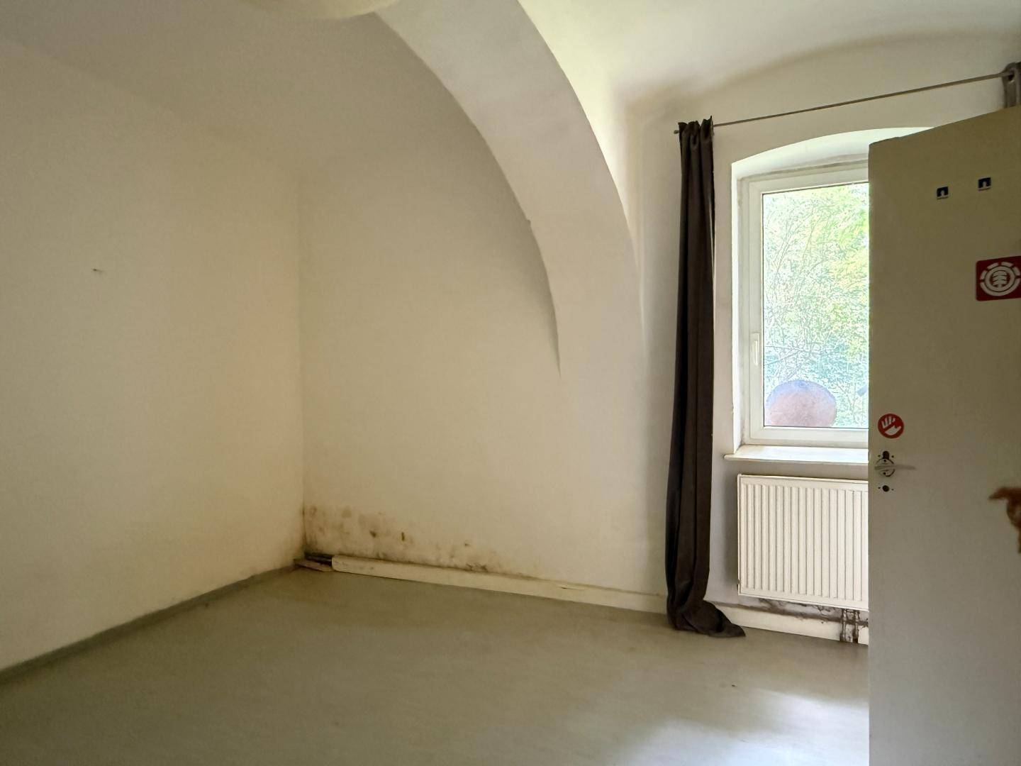 Einfacher Raum mit Fenster, Heizkörper und gewölbter Decke, der Renovierungsbedarf aufweist.