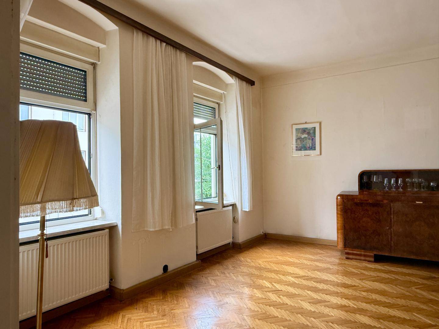 Heller Raum mit Fischgrätparkett, einem Fenster mit Vorhängen und einem antiken Sideboard.