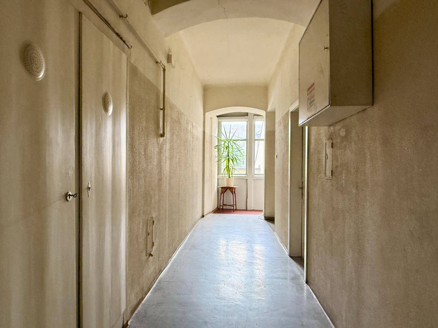 Langer, schmaler Flur mit hellen Wänden, Türen und einem Fenster am Ende, das Tageslicht hereinlässt.