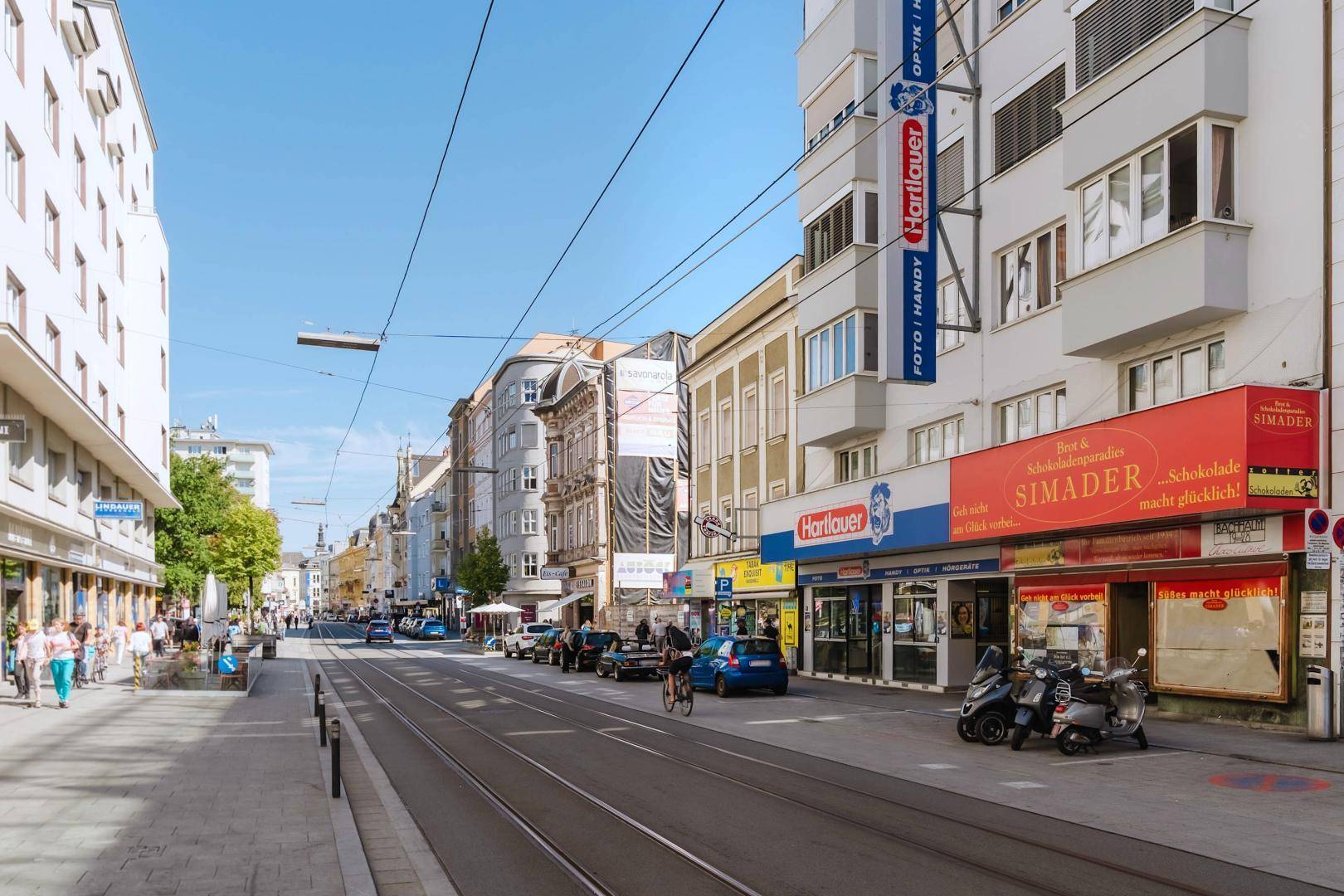 Eine belebte Stadtstraße an einem sonnigen Tag mit Geschäften, Gebäuden und öffentlichen Verkehrsmitteln.