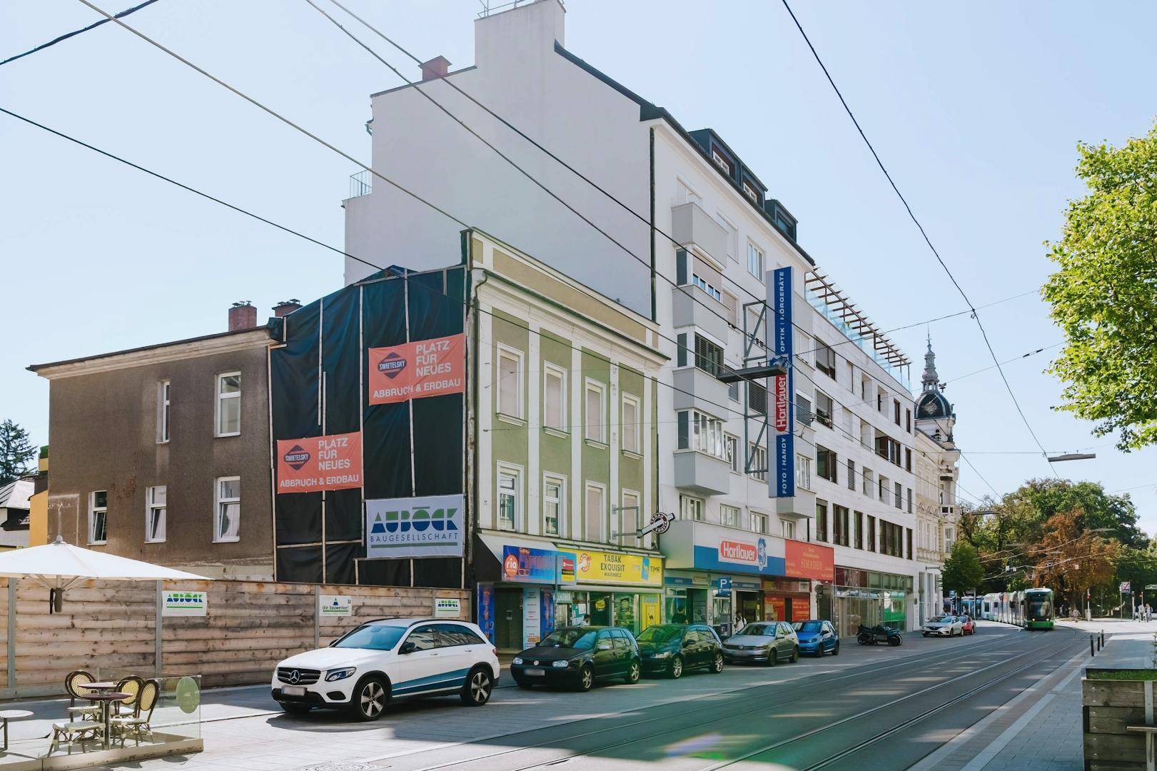 Stadtansicht mit Gebäuden, von denen einige renoviert werden, und einer belebten Straße mit parkenden Autos.