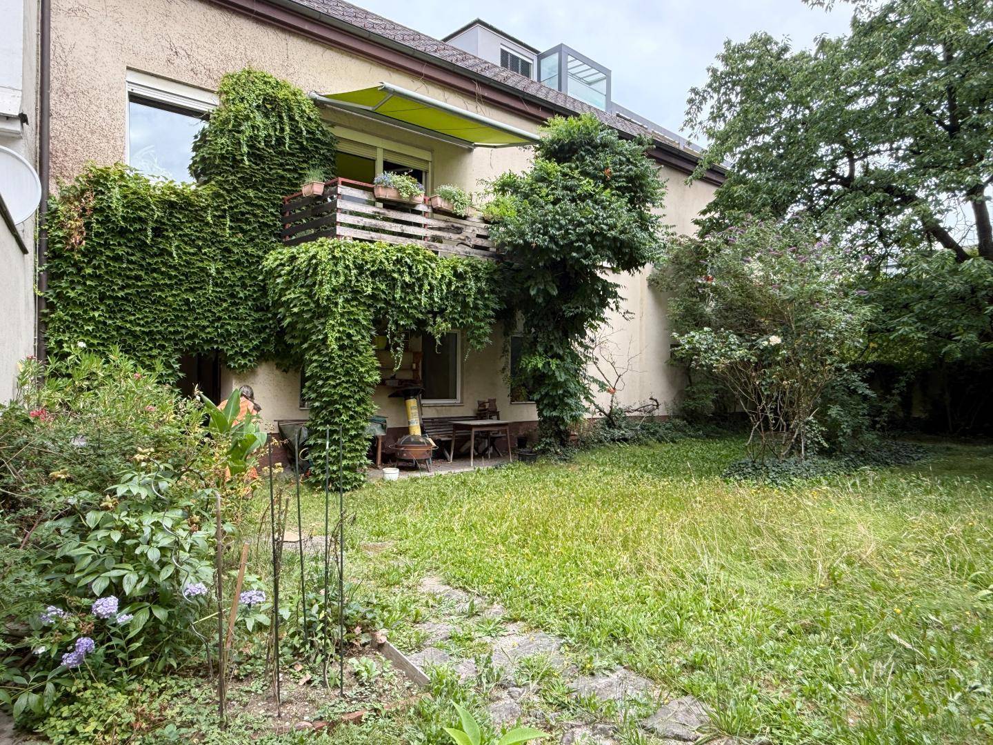 Gartenbereich mit Rasen und Pflanzen vor einem Gebäude mit Balkon, der von Kletterpflanzen umrankt ist.