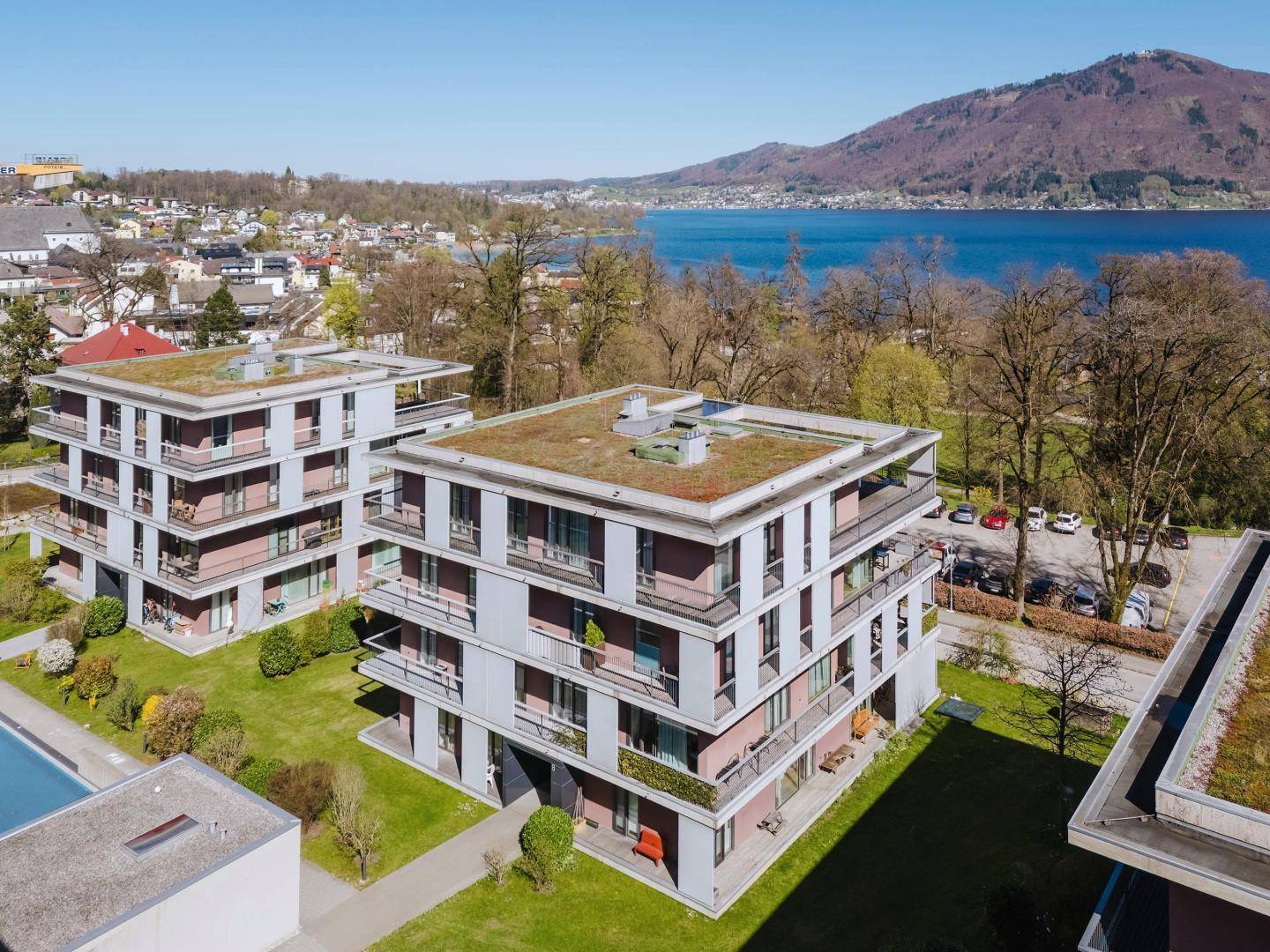 Wohnanlage mit modernen Gebäuden, Gründächern und einem See im Hintergrund unter blauem Himmel.