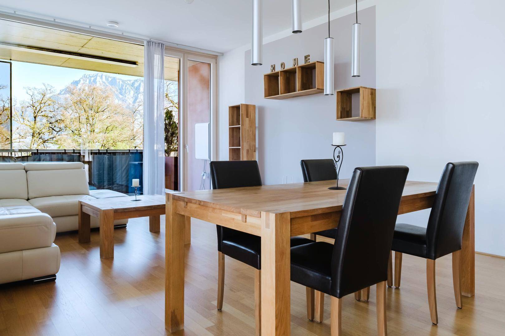 Heller Essbereich mit Holztisch, schwarzen Stühlen und Zugang zum Balkon mit Bergblick.