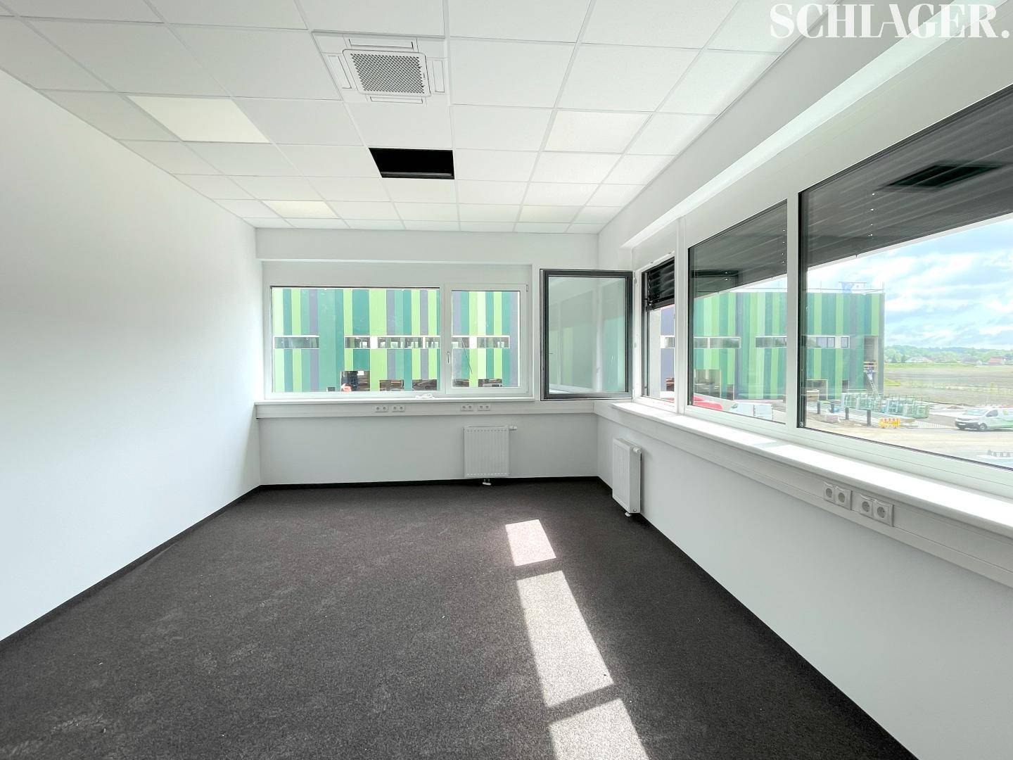 Leeres, helles Büro mit Teppichboden, großen Fenstern und Blick auf die grüne Fassade des Nachbargebäudes.