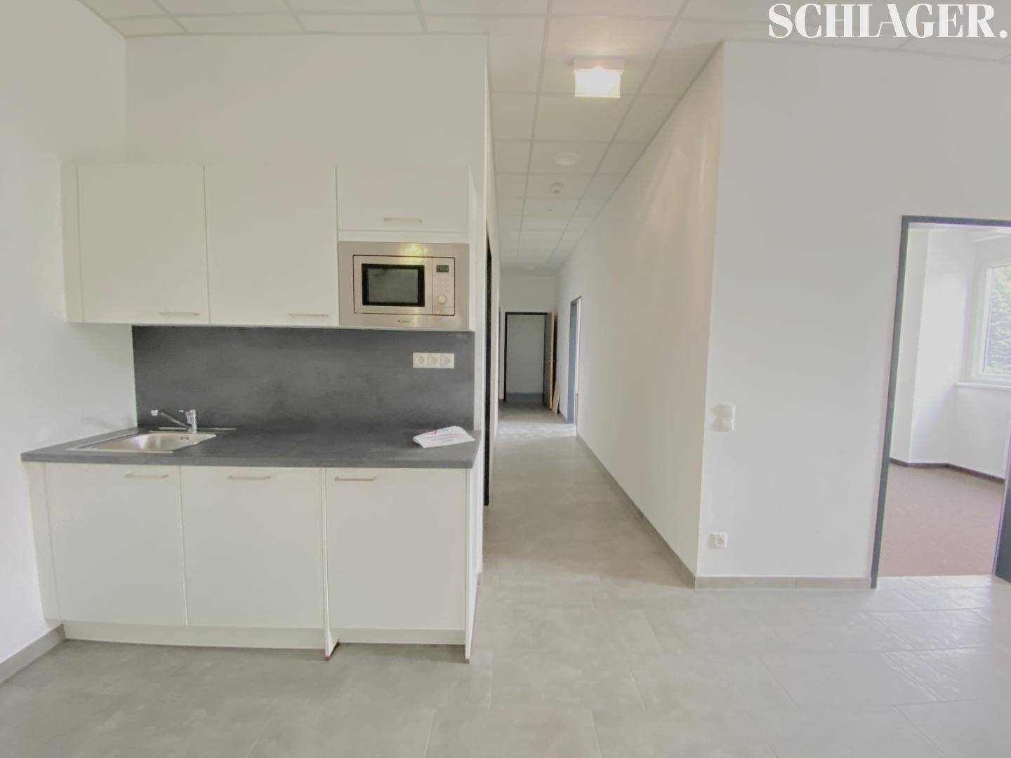 Kleine, moderne Küchenzeile mit weißen Schränken, integrierter Mikrowelle und Fliesenboden in einem Bürobereich.