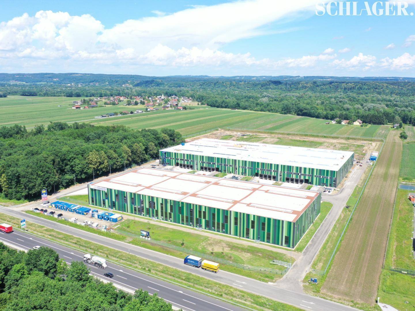 Weitläufige Luftaufnahme von zwei modernen Industriegebäuden mit grünen Fassaden in ländlicher Umgebung neben einer Autobahn.